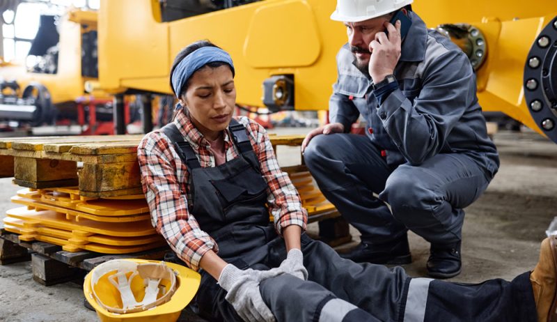 Young female engineer with hurting knee sitting by man calling ambulance Young female engineer with hurting knee sitting on the floor by anxious male worker in safety helmet and uniform calling ambulance