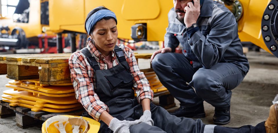 Young female engineer with hurting knee sitting on the floor by anxious male worker in safety helmet and uniform calling ambulance